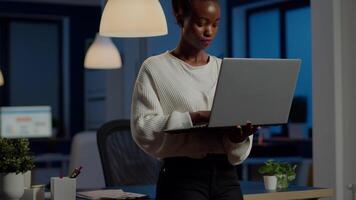 Black business woman looking at camera smiling holding laptop standing near desk in start-up company late at night. Focused employee using technology network wireless doing overtime for job video