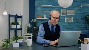 Senior old man listening music on headphones while working on laptop in living room and wife drinking coffee in background. Retired man using modern technology reading typing, searching on computer video