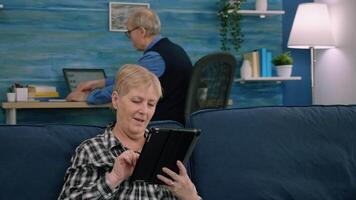 Senior woman reading emails on digital tablet sitting on sofa at home, while senior adult man working at laptop in background. Person enjoying using notepad browsing internet shopping video