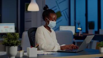 African employee with protection face mask working at laptop overtime in new normal business office to respect deadline of project, analysing documents sitting at desk overtime during global pandemic video