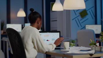 Busy african business woman analysing financial reports checking company statistics graphs, looking at laptop, pointing to numbers late at night in start-up office doing overtime to respect deadline video
