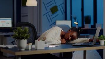 Exhausted overload african business woman falling asleep on desk with open laptop monitor while working in start up company. Overworked employee using doing overtime respecting deadline, sleeping video