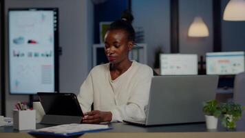 African manager woman using laptop and tablet in same time working overtime in business start-up office. Busy multitasking Busy employee analysing financial statistics overworking writing, searching. video