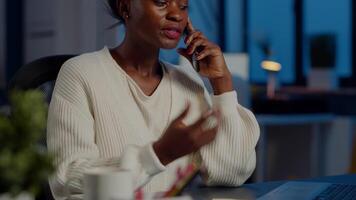 Nervous african manager discussing at smartphone with employee working overtime sitting at desk in business office late at night resolving financial problem. Furious businesswoman overworking video