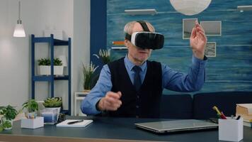 Senior man using virtual reality goggles in living room gesturing sitting in home workplace. Retired man experiencing vr headset while woman watching tv sitting on sofa in background. video