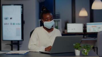 African woman with face mask reading emails late at night to respect deadline of project working in new normal business office, analysing documents, making strategy overtime during global pandemic video