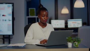 Stressed african manager woman working with financial graphs checking graphs, typing on laptop, reading raports late at night in start-up office doing overtime to respect deadline of project. video