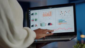 Back view of african business woman analysing financial statistics holding laptop standing in start-up office overworking. Busy focused employee using modern technology network wireless overtime video