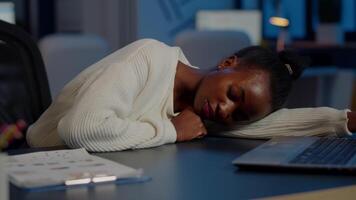 Dark skin freelancer doing overtime falling asleep with hand on desk trying to respect deadline for project. African employee working late at financial project sleeping on table in strat up office video