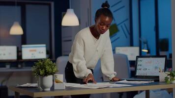 Stressed african manager woman working with financial documents standing at desk checking graphs, holding papers, reading raports late at night in start-up office doing overtime to respect deadline. video