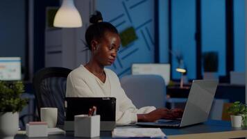 Multitasking black business woman working at laptop and tablet in same time doing overtime in start-up office. Busy african employee analysing financial statistics overworking writing, searching. video