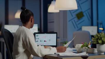 African business woman holding documents with graphics and financial statistics working overtime in front of laptop sitting in start-up business office. Focused employee respecting deadline of project video