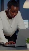 African business woman standing near desk looking at camera after reading mails on laptop working in start-up company late at night. Focused employee using technology network wireless doing overtime video