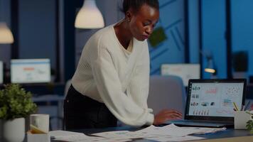 Stressed african manager woman working with financial documents checking graphs, holding papers, reading raports, standing at desk late at night in start-up office doing overtime to respect deadline. video