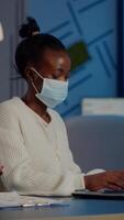 African employee with protection face mask working at laptop overtime in new normal business office to respect deadline of project, analysing documents sitting at desk overtime during global pandemic video