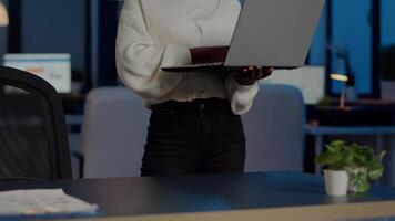 Happy african freelancer looking at camera smiling holding laptop standing near desk in start-up company late at night. Focused employee doing overtime for job respecting deadline of financial project video