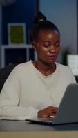 Stressed african manager woman working with financial graphs checking graphs, typing on laptop, reading raports late at night in start-up office doing overtime to respect deadline of project. video