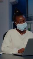 African woman with face mask reading emails late at night to respect deadline of project working in new normal business office, analysing documents, making strategy overtime during global pandemic video
