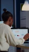 Busy african business woman analysing financial reports checking company statistics graphs, looking at laptop, pointing to numbers late at night in start-up office doing overtime to respect deadline video