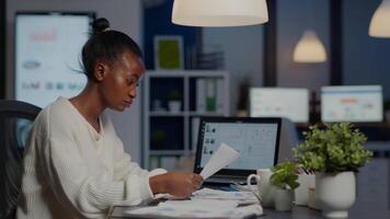 Black manager analysing graphics and financial statistics from documents working overtime in front of laptop sitting in start-up business office. Focused employee respecting deadline of project video