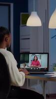 African freelancer working remotely discussing with woman partner online sitting in front of laptop in start-up office talking during virtual meeting at midnight, using headphone doing overtime video