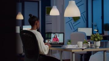 African freelancer working remotely discussing with woman partner online sitting in front of laptop in start-up office talking during virtual meeting at midnight, using headphone doing overtime video