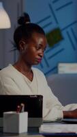 Multitasking black business woman working at laptop and tablet in same time doing overtime in start-up office. Busy african employee analysing financial statistics overworking writing, searching. video