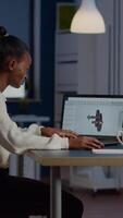 African engineer working late at night on a 3D model of industrial gears at laptop doing overtime in start up office. Freelancer studying prototype idea on pc showing cad software on device display video
