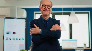 Elderly successful businessman entrepreneur standing looking at camera crossing arms and smiling. Manager working in professional start up financial business, modern company workplace ready for meeting video