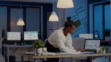 Black bookkeeper working on financial reports checking statistics graphs, looking at laptop, pointing to numbers standing at desk late at night in start-up office doing overtime to respect deadline video