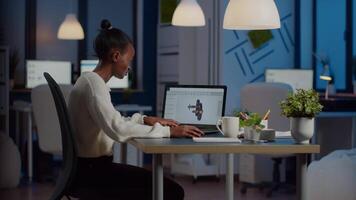 African engineer working late at night on a 3D model of industrial gears at laptop doing overtime in start up office. Freelancer studying prototype idea on pc showing cad software on device display video
