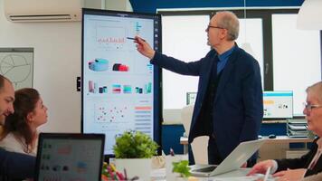 Mature CEO CTO executive director presenting statistical data, financial project briefing diverse group of employees. Multiethnical team working in professional startup company during conference meeting video