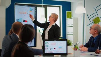 Elderly project manager pointing at desktop presenting statistical data, briefing diverse group of employees. Multiethnical team working in professional startup financial company during conference video