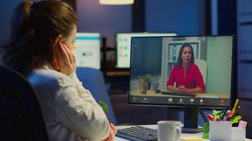 Freelancer working remotely discussing with woman partner online sitting in front of computer in start-up office talking during virtual meeting at midnight, using wireless headphone doing overtime video