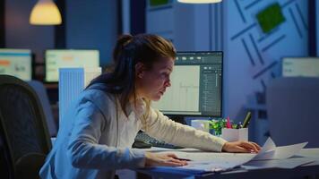 Focused woman architect checking and matching blueprints sitting at desk in front of computer late at night. Designer using arhitecture plans of buildings working overtime, creating and studying video