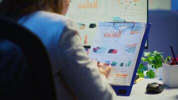 Close up of business woman holding clipboard with graphics and financial statistics working overtime in front of computer sitting in start-up business office. Busy employee using modern technology video