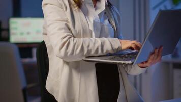Freelancer woman holding laptop, writting and looking at camera smiling standing in start-up company late at night. Focused employee using technology network wireless doing overtime for job reading. video
