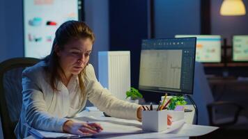 Overworked woman architect analysing and matching blueprints sitting at desk in front of computer late at night. Designer using arhitecture plans of buildings working overtime, creating and studying video