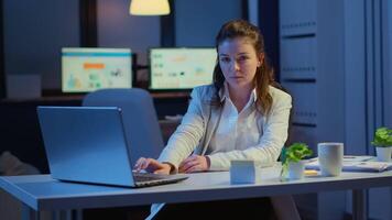 Business woman looking at camera smiling after typing on laptop sitting at desk in start-up company late at night. Focused employee using technology network wireless doing overtime for job reading. video