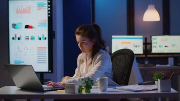 Business woman working overtime checking team project, taking notes, drinking coffee, analysing financial documents sitting at desk in start-up company late at night using modern technology network video