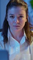 Vertical . Close up portrait of business woman smiling at camera after reading mails on laptop sitting at desk in start-up company late at night. Focused employee using technology network wireless doing overtime video