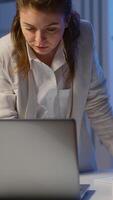 Vertical . Focused tired business woman looking at camera after reading mails on laptop standing near desk in start-up company late at night. Focused employee using technology network wireless doing overtime video