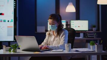 Manager woman with face mask working overtime in new normal business office checking team project, taking notes, analysing financial documents sitting at desk late at night during global pandemic video