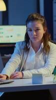 Vertical . Business woman looking at camera smiling after typing on laptop sitting at desk in start-up company late at night. Focused employee using technology network wireless doing overtime for job reading. video