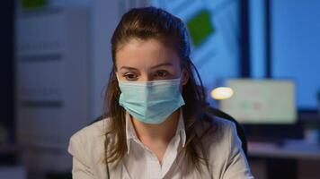 Close up of woman with face mask reading emails late at night to respect deadline of project working in new normal business office, taking notes, analysing documents overtime during global pandemic video