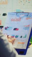 Vertical . Close up of business woman holding clipboard with graphics and financial statistics working overtime in front of computer sitting in start-up business office. Busy employee using modern technology video
