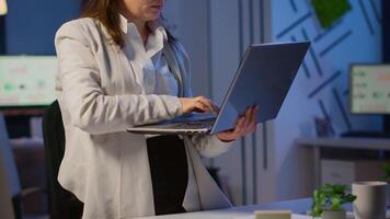 Exhausted business woman holding laptop, typing and sighing looking at camera standing in start-up company late at night. Employee using technology network wireless doing overtime for job reading. video