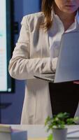 Vertical . Tired manager raising head looking at camera sighing holding laptop typing on it, working late at night in start-up business office. Focused employee using technology network wireless doing overtime video