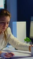 Vertical . Overworked woman architect analysing and matching blueprints sitting at desk in front of computer late at night. Designer using arhitecture plans of buildings working overtime, creating and studying video