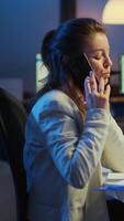 Vertical . Nervous manager discussing at smartphone with employee working overtime sitting at desk in business office late at night resolving financial problems. Busy employee using modern technology network video